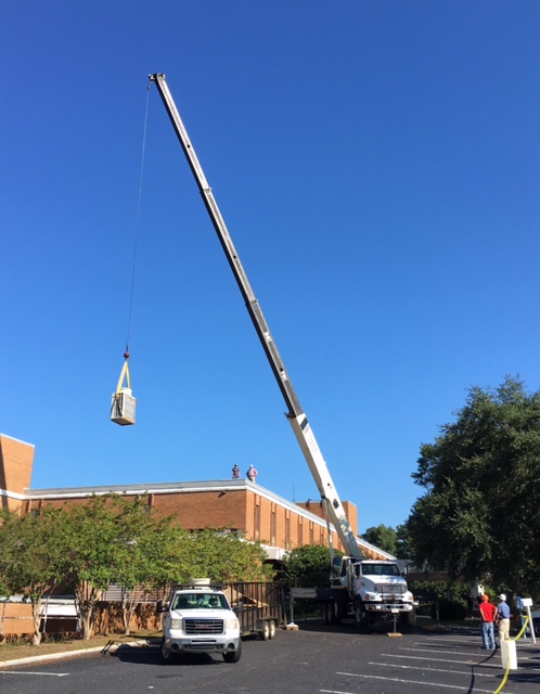 A crane is lifting an AC unit up and over DMC rooftop to be installed.